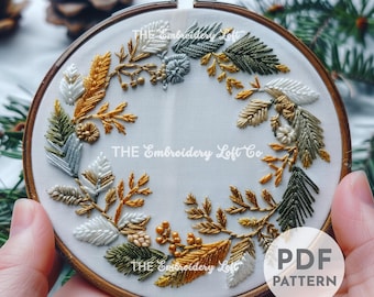 May include: A white embroidery hoop featuring a gold, green and white floral wreath design. The text "THE Embroidery Loft Co" is visible in the centre of the hoop. The hoop is being held by a hand. The text "PDF PATTERN" is visible in the bottom right corner of the image.