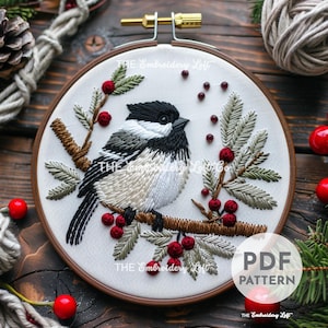 May include: A white embroidery hoop with a black and white chickadee bird perched on a branch with red berries and green leaves. The hoop is on a wooden surface. The text "THE Embroidery Loft" is printed on the hoop.
