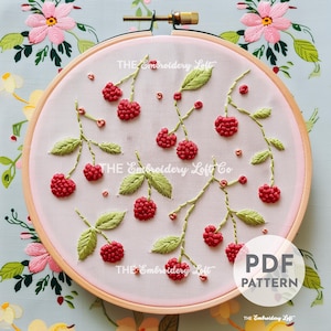 Könnte beinhalten: Ein rosa Stickrahmen mit einem Design aus grünen Blättern und roten Beeren. Der Text "THE Embroidery Loft Co" ist in der Mitte des Rahmens sichtbar. Der Rahmen befindet sich auf einem blauen und weißen Blumenhintergrund.