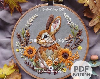 Puede incluir: Arte de bordado en aro con un conejo marrón con una corona floral de girasoles, hojas y bayas. El texto "THE Embroidery Loft Co" es visible en la corona.