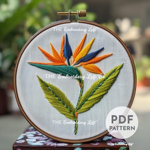 Puede incluir: Flor de ave del paraíso bordada en un aro de bordado blanco con hojas verdes y un marco de madera. El texto "THE Embroidery Loft" es visible en el aro.