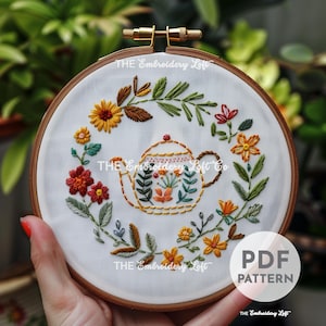 Puede incluir: Patrón de bordado para una tetera rodeada de una corona de flores. El patrón está en un aro e incluye el texto "THE Embroidery Loft Co."
