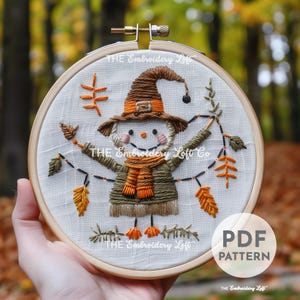 May include: Embroidered hoop art featuring a whimsical snowman wearing a witch hat and scarf, surrounded by autumn leaves and a string of lights. The design is stitched on a white linen fabric and framed in a wooden hoop. The text "THE Embroidery Loft" appears at the top and bottom of the hoop.
