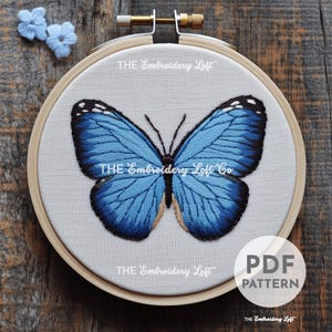 Op de afbeelding: Een ronde houten borduurring met een stoffen paneel met een handgeborduurd blauw vlinderontwerp. De vlinder heeft tinten licht- en donkerblauw met zwarte vleugelranden. De tekst THE EMBROIDERY LOFT is zichtbaar op de stof.