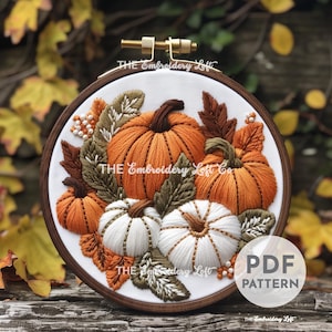 以下が含まれることがあります： オレンジと白の色合いのカボチャとひょうたんの集まりが、緑の葉に囲まれた刺繍のフープアート。フープは木でできており、白い布の背景があります。右下隅に「PDF PATTERN」の文字が見えます。