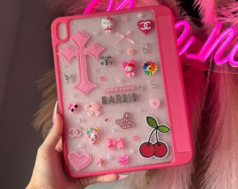 May include: A pink iPad case decorated with various charms, including a cross, hearts, bows, and a cherry sticker. The word "BARBIE" is spelled out in silver letters, and the word "HARJUKU" is above it.