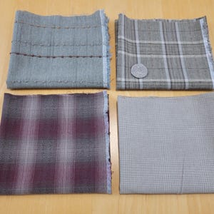 May include: Four fabric swatches in shades of grey, green, and burgundy are arranged on a light wood surface. The swatches display various patterns, including stripes, plaid, and a grid design. A coin is placed on one of the plaid swatches.