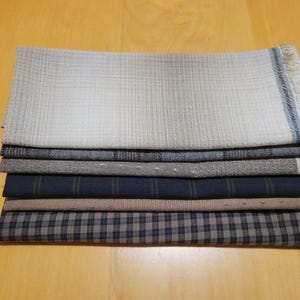 May include: A pile of seven folded fabric cloths in different patterns and colours. The top cloth is light beige with a grid pattern and a blue stripe along the edge. Other cloths include grey, navy, and brown with dotted, striped, and checkered designs.