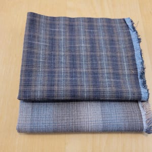 May include: Two folded fabric pieces with a plaid pattern. The top piece is navy blue and brown, while the bottom piece is grey and blue. The edges of the top piece are frayed.