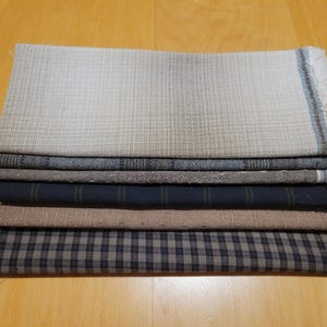 May include: A collection of folded fabric textiles in a variety of patterns and colours. The textiles include a light beige plaid, a dark navy blue with thin stripes, and a brown and navy blue checkered pattern. The fabrics are stacked on a wooden surface.