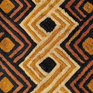 May include: A textured rug featuring a repeating geometric pattern of nested diamonds. The design consists of thick, raised lines in alternating colours of black, deep orange, mustard yellow, and cream, creating a bold, high-contrast visual effect.