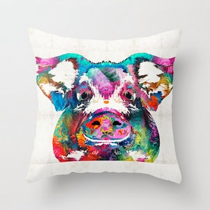 May include: Square throw pillow featuring a colorful, abstract painting of a pig's face. The pig's snout, ears, and eyes are depicted with vibrant hues of blue, pink, green, and orange. The pillow has a white background.