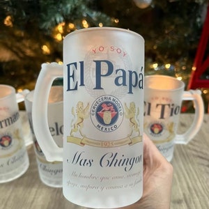 May include: A frosted glass beer stein with a white background and a blue and gold design. The stein features the text "Yo Soy El Papa" and a logo for Cerveceria Modelo. The text "Mas Chingon" is below the logo, and the text "Un hombre que ama, inspira y anima a su familia" is below that.