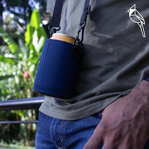 May include: A navy blue can cooler with a black strap is worn over the shoulder. The cooler is holding a gold and yellow can. The person is wearing a green shirt and blue jeans. A white bird logo is on the shirt.