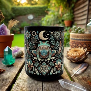 May include: Black ceramic mug featuring a detailed illustration of a frog with teal and brown accents, surrounded by celestial and floral designs. The mug has a wide, cylindrical shape, perfect for coffee or tea.