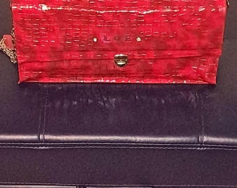 May include: A red crocodile embossed clutch purse with silver studs and a silver clasp. The purse has the word "LOVE" embossed on the front.