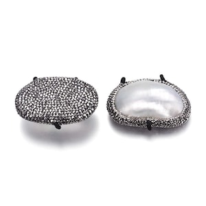 May include: Two oval-shaped jewelry pieces. One is covered in small, dark gray stones, and the other features a large, lustrous white pearl surrounded by a border of dark gray stones. Both have black attachments.