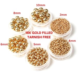 以下が含まれることがあります： 2mmから10mmまでの様々なサイズの金色のビーズが入った6つの透明な容器。中央には「18K GOLD FILLED TARNISH FREE」の文字。これらのビーズは、ジュエリー作りや工芸に適しています。
