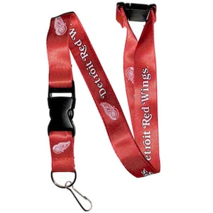 May include: Red lanyard with the text "Detroit Red Wings" and a logo. The lanyard has a black plastic clasp and a metal clip. The lanyard is red with white text and logo.