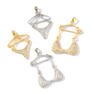 May include: Four metal pendants shaped like miniature clothing hangers with attached bikini bottoms or tops, encrusted with small clear crystals. Two pendants are gold-toned and two are silver-toned, each featuring a small loop at the top.