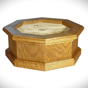 May include: An octagonal wooden box with a light brown finish. The box has a multi-tiered design with a lighter-colored interior. The wood grain is visible, adding texture to the piece. The box is likely used for storage or display.