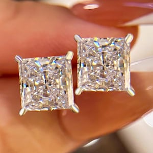 May include: A pair of square-cut diamond stud earrings. The earrings are set in a silver metal and feature a radiant, multi-faceted design. The diamonds sparkle with a brilliant, clear light, showcasing their intricate cuts. These earrings are a classic jewelry piece.