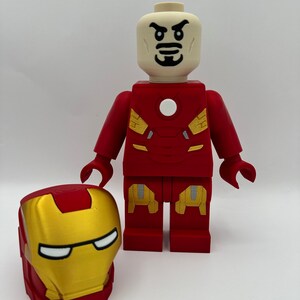 May include: A red and gold Lego minifigure of Iron Man with a stern expression. The minifigure is wearing a red and gold suit with a white circle on the chest. The minifigure is holding a separate red and gold Lego helmet.
