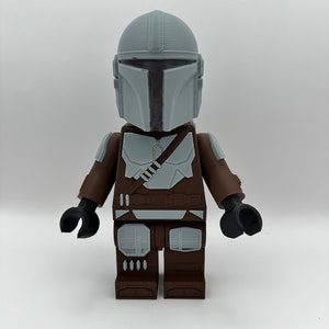 May include: A large, blocky figure of The Mandalorian, featuring a gray helmet and armor details. The figure has a brown torso and legs, with black arms and hands. The design is reminiscent of a popular building block toy.