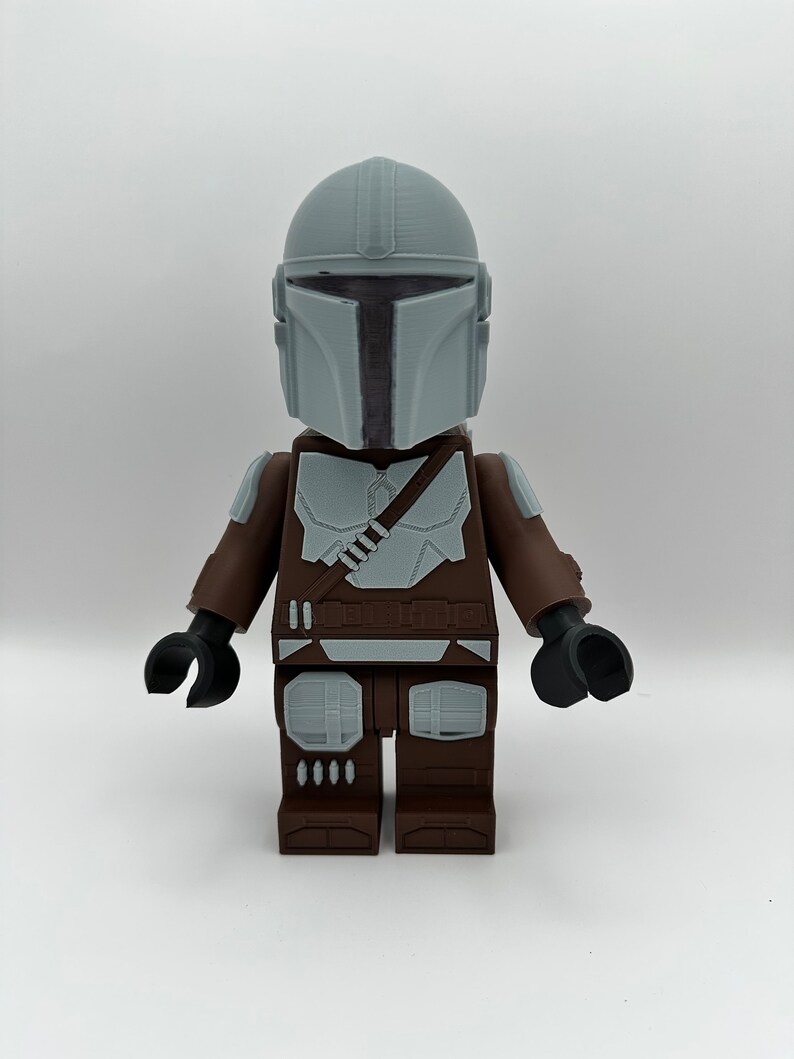 May include: A large, blocky figure of The Mandalorian, featuring a gray helmet and armor details. The figure has a brown torso and legs, with black arms and hands. The design is reminiscent of a popular building block toy.
