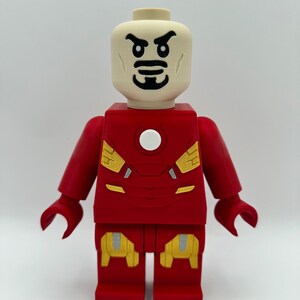 May include: A red and gold Lego minifigure of Iron Man. The minifigure has a white face with black eyes and a black mustache. The minifigure is wearing a red and gold suit with a white circle on the chest.