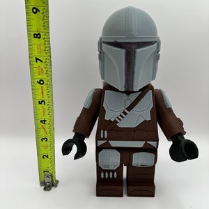 May include: A large, 3D-printed Mandalorian figure, approximately 9 inches tall, with a gray helmet and armor details. The figure has brown and gray accents, black hands, and a utility belt. The figure is a stylized representation of the character.