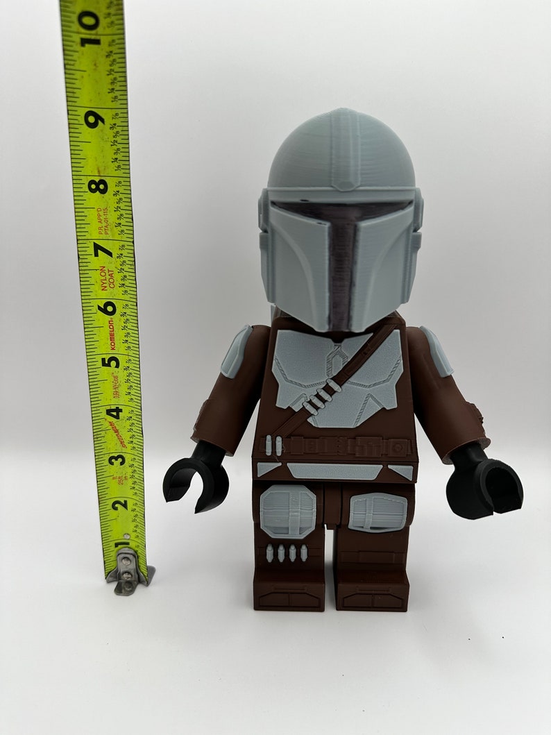 May include: A large, 3D-printed Mandalorian figure, approximately 9 inches tall, with a gray helmet and armor details. The figure has brown and gray accents, black hands, and a utility belt. The figure is a stylized representation of the character.