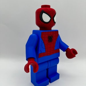 May include: A large, blue and red Spider-Man figure, resembling a Lego minifigure. The figure has a red head with white eyes and a black outline, and a red and blue torso with a black spider emblem. The hands are red.