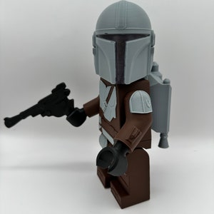 May include: A 3D-printed Mandalorian figure, featuring a gray helmet, brown armor, and a black blaster. The figure has a jetpack and is posed against a white background. The figure is a toy.