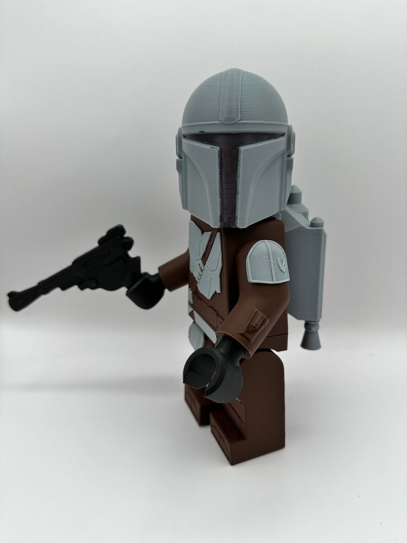 May include: A 3D-printed Mandalorian figure, featuring a gray helmet, brown armor, and a black blaster. The figure has a jetpack and is posed against a white background. The figure is a toy.