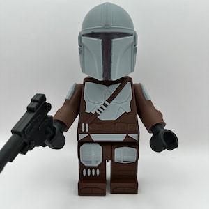 May include: A Mandalorian action figure, featuring a gray helmet and armor, brown arms and legs, and a black blaster. The figure is a block-style design, reminiscent of a popular building toy.