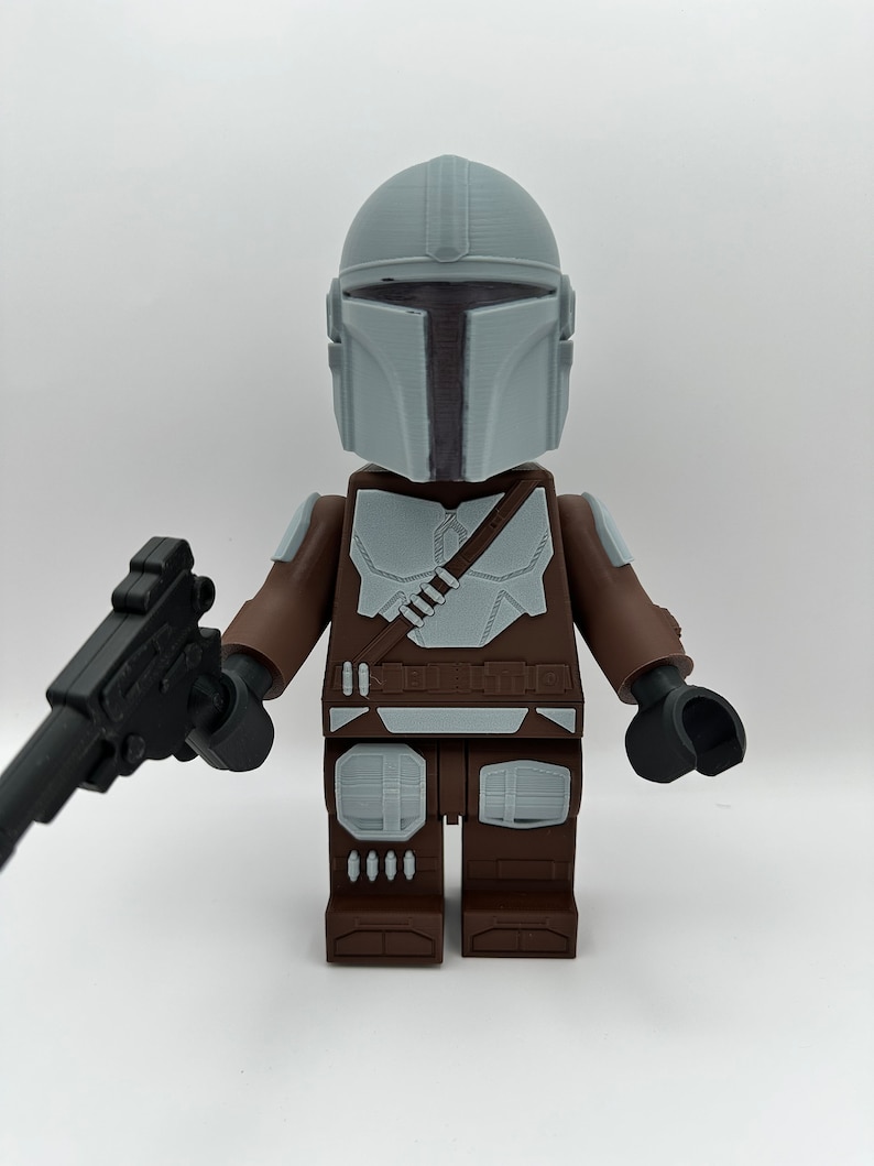 May include: A Mandalorian action figure, featuring a gray helmet and armor, brown arms and legs, and a black blaster. The figure is a block-style design, reminiscent of a popular building toy.