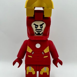 May include: A red and yellow Lego minifigure of Iron Man with a white face and a black mustache. The minifigure has a yellow helmet with a red visor and a red chest plate with a white circle in the center.