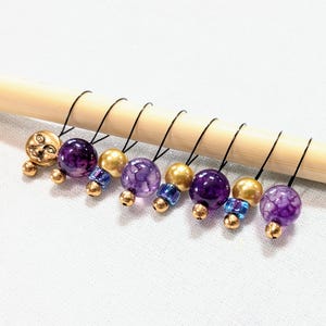 May include: A collection of handmade stitch markers. The markers feature a variety of beads, including gold, purple, and blue, attached to black metal hooks. One marker has a gold face charm.