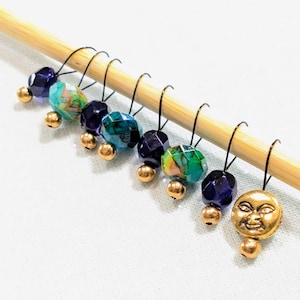 May include: A collection of colorful stitch markers on a wooden knitting needle. The markers feature faceted beads in shades of blue, green, and purple, with gold-colored accents. One marker has a gold-colored smiling moon charm.