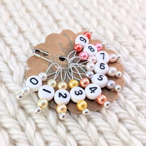 Könnte beinhalten: Ein Set mit zehn bunten Maschenmarkierern mit den Zahlen 0-9. Die Markierer sind an einer braunen Karte mit einer Sicherheitsnadel befestigt. Die Markierer sind aus weißem Kunststoff mit schwarzen Zahlen und haben an jedem Ende eine Perle.