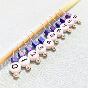 May include: A set of ten numbered stitch markers. Each marker has a white bead with a black number, a silver bead, and a purple square bead. The markers are on a wooden knitting needle.
