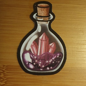 Könnte beinhalten: Eine Glasflasche mit einem Korkverschluss, gefüllt mit rosa Kristallen und einer lila, glitzernden Flüssigkeit.