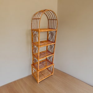 Puede incluir: Una estantería alta de mimbre tejido con forma de arco y cinco estantes. La estantería está hecha de mimbre marrón natural y tiene un tinte marrón claro.
