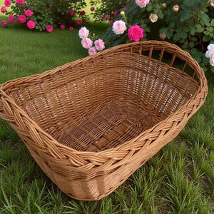 Puede incluir: Una cesta de mimbre tejida de color marrón claro, de forma rectangular con lados curvos. La cesta está sobre hierba verde, con rosas rosas en el fondo. La cesta está hecha de material tejido.