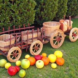 Puede incluir: Un tractor decorativo de madera con componentes de cesta tejida y ruedas. El tractor es marrón y beige, y está rodeado de una variedad de frutas coloridas, incluyendo manzanas, naranjas y limones. La escena está ambientada en un césped verde.
