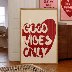May include: A white framed poster with a red heart shape containing the text "GOOD VIBES ONLY" in white. The poster is hanging on a wall with two other posters, one with the text "IT'S OKEY IF YOU DON'T LIKE ME NOT EVERYONE" and the other with the text "THIS IS MY HAPPY PLACE" with a red heart.