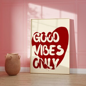 May include: A white framed poster with a red heart shape and the text "GOOD VIBES ONLY" in white lettering. The poster is hanging on a pink wall.