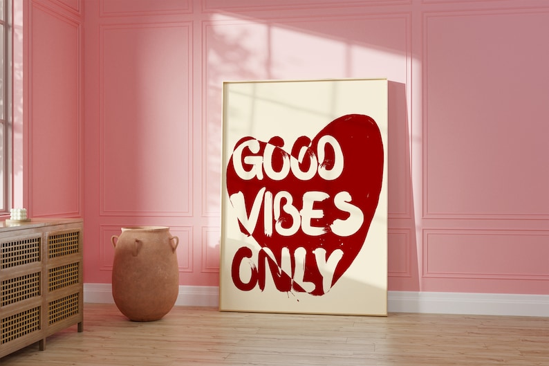 May include: A white framed poster with a red heart shape and the text "GOOD VIBES ONLY" in white lettering. The poster is hanging on a pink wall.