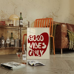 May include: A white framed print with a red heart shape and the text "GOOD VIBES ONLY" in white. The print is leaning against a wooden shelf with bottles and glasses on it. There is a brown leather couch and an orange metal chair in the background.